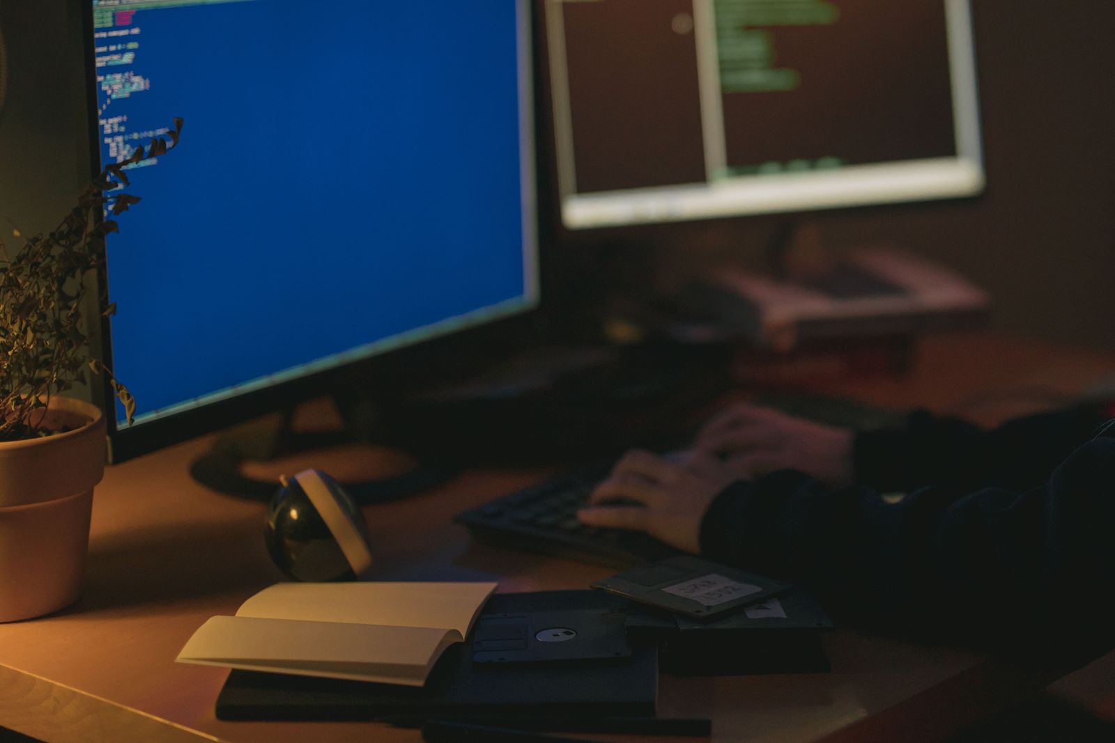 A late-night desk lit by two computer screens, with a keyboard, a plant and an open notebook